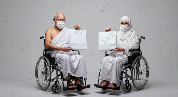 A man and a woman are sitting in wheelchairs, both wearing white Ihram clothing. The man is in traditional male Ihram attire while the woman is covered with an abaya and hijab. Each is holding a white bag and wearing medical face masks. They appear to be in a neutral grey studio environment.