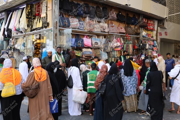 An image of a group of people gathering in front of a shop selling bags and various products. Some individuals are wearing white ihram clothing, while women are wearing black abayas and hijabs. The shop is filled with hanging bags and fabrics displayed in the storefront. The scene suggests a busy local market, possibly located in Saudi Arabia.