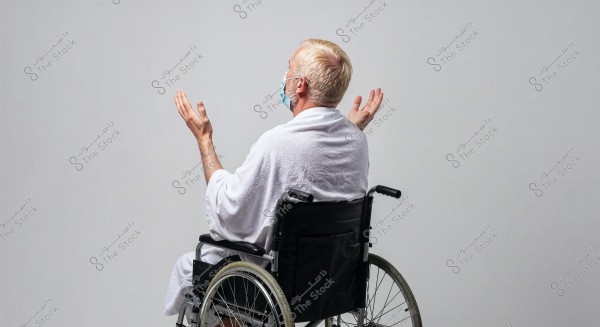A man sitting in a wheelchair wearing a white robe and a medical mask on his face. The man has his hands slightly raised in a gesture resembling prayer or supplication. The background is plain white.