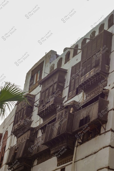 An image of a heritage building featuring traditional wooden windows (Rawashin) with ornate balconies protruding from the walls. The building is painted white and shows signs of aging, highlighting the traditional architectural style found in a historical area. A green palm leaf appears on the side, adding a natural touch.