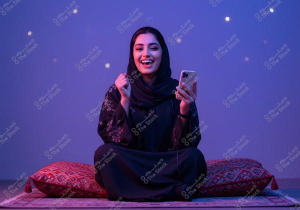 An image of a woman wearing a decorated black abaya and hijab, sitting on a cushion with traditional patterns, holding a smartphone. She appears smiling and happy. The room is lit with soft blue lighting, with small light spots in the background.
