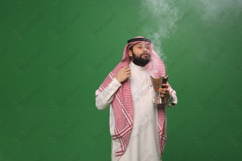 رجل سعودي يقف على خلفية خضراء يرتدي الثوب و الشماغ، يقوم بعمل حركات بيديه ، يبخر نفسه بالطيب و العود، احتفالات العيد