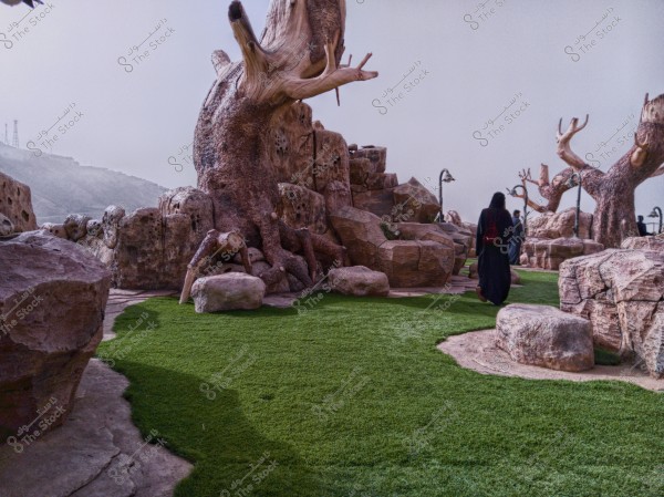 An image of a natural scene featuring a large, branching tree on a landscape covered with artificial green grass. Surrounding the tree are large, variously shaped rocks. In the background, there are misty hills with several aerial towers. A person in dark clothing is walking away from the camera.