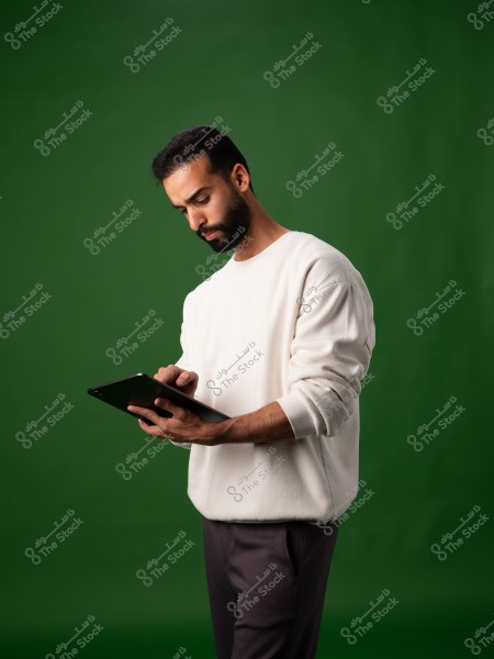 A portrait of a man holding and looking at a tablet with focus. He is wearing a white long-sleeved shirt and dark pants. The background is green, which highlights the individual clearly.