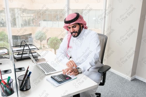 رجل سعودي يرتدي الزي السعودي التقليدي يعمل في مكتب بواجهة زجاجية ويستخدم قلم رصاص لتدوين الملاحظات