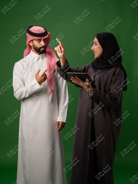 رجل يرتدي الثوب السعودي التقليدي يتواصل مع امرأة ترتدي الحجاب وتحمل جهازًا لوحيًا أمام خلفية خضراء.