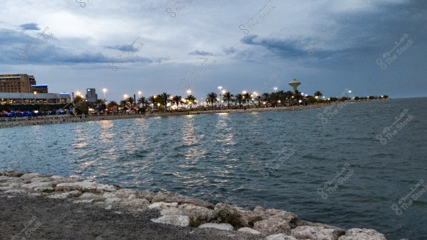 The image depicts a scenic view of a waterfront bay with a cloudy sky. There is a paved area on the left and a shoreline extending to the right. Illuminated buildings and palm trees line the coast, with streetlights adding vibrancy to the scene. A water tower is visible in the background.