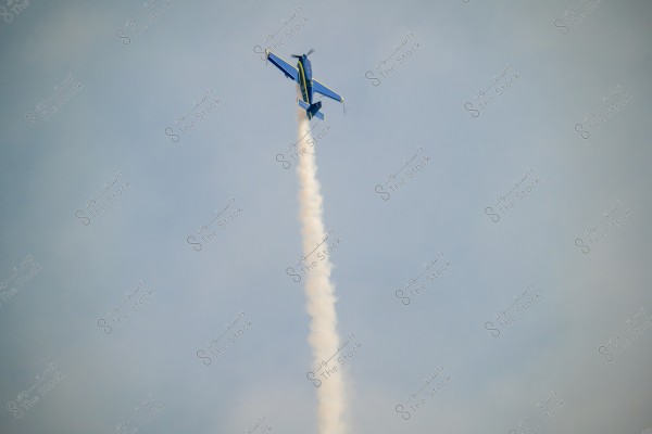 A blue aircraft performing an aerial display, flying vertically in the sky. A trail of white smoke follows the plane. The sky is clear, and the aircraft appears to be slightly inverted.