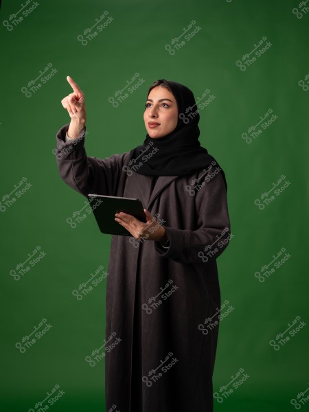 The image shows a woman wearing a black abaya and hijab, standing against a green background. She is holding a tablet in one hand and pointing forward with her finger, giving the impression that she is interacting with something invisible.
