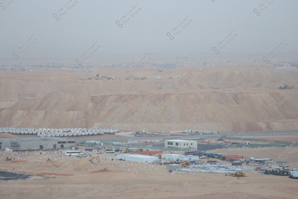 A general view of a construction site in a desert area, featuring a collection of temporary buildings and parked heavy construction equipment. The area is surrounded by sandy hills, with a hazy skyline in the background.