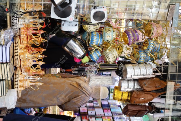 An image of a shop selling various traditional goods. The picture includes a collection of intricately designed golden vessels with an Eastern traditional style, several mugs and plates decorated in gold and blue, some small electronic devices, brown and silver bags, and several leather items. In the background, shelves display books and various items. A person in the background appears to be wearing traditional clothing similar to a thobe and shemagh.