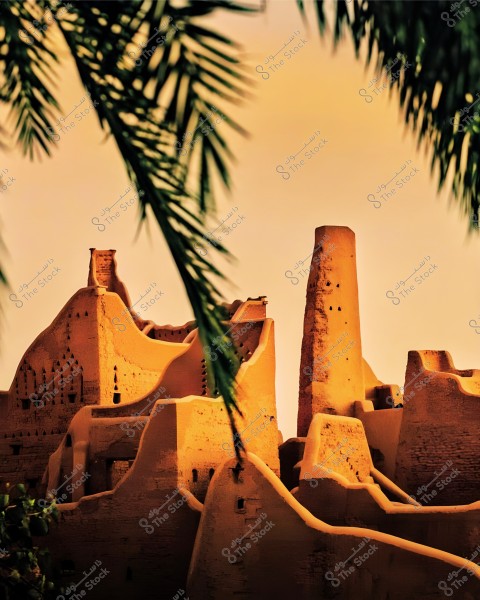An image depicting traditional mud buildings in the open air, showcasing ancient castle-like architectural structures bathed in golden sunlight. Green palm leaves are prominent in the foreground, providing a natural contrast to the warm color of the structures. The sky in the background is a deep orange hue, creating a serene and beautiful atmosphere.