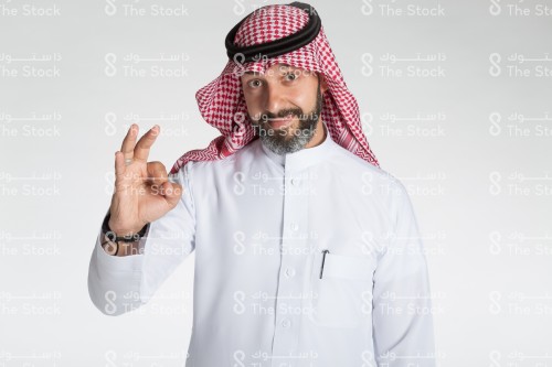 بورتريه رجل سعودي يقف على خلفية بيضاء، يرتدي الزي السعودي الرسمي، ينظر الى الكاميرا ويقوم بعمل حركات بيديه، تعبير على اتمام شيء او الاتفاق مع شيء