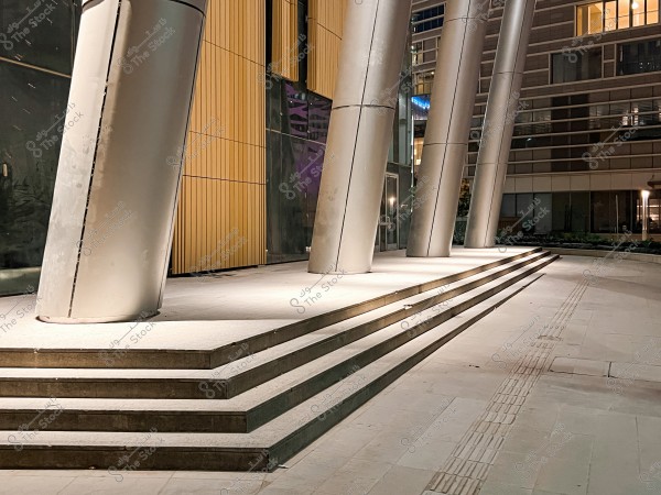 An image depicting the entrance of a modern building with large tilted metal columns and wide concrete steps. The walls are clad with large wooden panels and in the background, there is a glass facade and parts of another building. The area is illuminated by night lights, giving it a contemporary appearance.