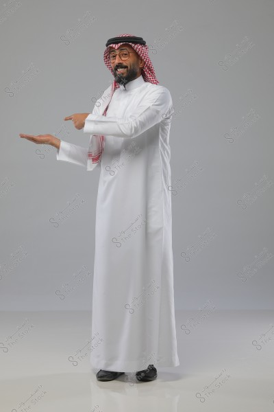 صورة لرجل يقف مبتسمًا ويرتدي الثوب الأبيض التقليدي والغترة الحمراء والشماغ والعقال الأسود, يشير بيده إلى الجانب بطريقة توحي بالتقديم أو التوضيح, وهو يقف في استوديو ذو خلفية رمادية.