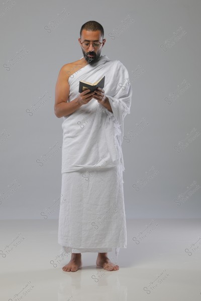 A bearded man wearing a white Ihram, standing and reading from a small book. The man is wearing glasses and has a serious expression. The Ihram consists of two white pieces of cloth, and he is standing on a white floor with a simple gray background.