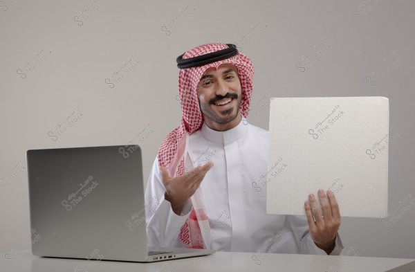 Image of a man sitting at a desk in front of a laptop, wearing traditional Saudi attire with a red and white checkered shemagh and an agal. The man is holding a large white board and smiling.