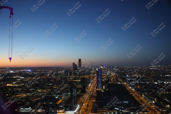 The image shows an aerial view of Riyadh city in Saudi Arabia during twilight. The sky displays a gradient of colors from orange to dark blue. Illuminated buildings with blue and white lights are visible, especially along the major roads, with a crane visible in the top left corner of the image.