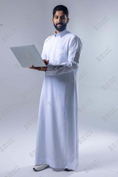 Image of a man standing while holding an open laptop. The man is wearing a traditional white thobe and appears to be smiling. The background is a neutral light gray, emphasizing the person in the image.