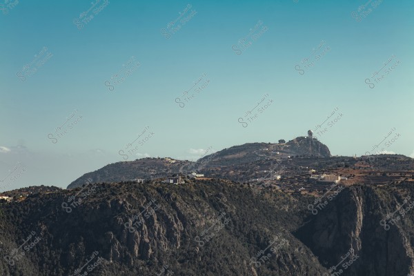 A panoramic image of a mountainous landscape, featuring lush green hills covered with trees in the foreground. In the background, there is a small cluster of buildings and terraced agricultural fields on the mountainside, with a large dome on top of the mountain under a clear blue sky.