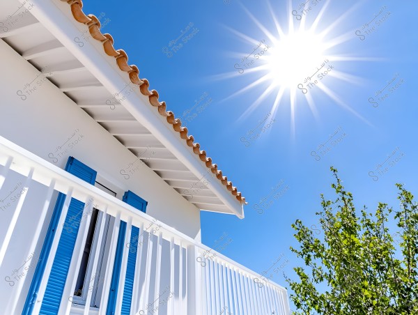 Image of a Mediterranean-style house featuring part of a roof with brown tiles against a clear blue sky. There is a window with a white frame and dark blue shutters. A white railing is in the foreground, with the bright sun shining at the top right of the image, and a branch of a green tree appears below.
