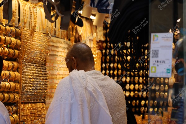 An image of a man wearing a white Ihram in a gold market. The man is seen from behind amidst a visual display of numerous gold jewelry, rings, and bracelets on the walls. A glass sign with stickers is visible on the right side.