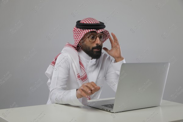 صورة لرجل يرتدي زيّاً تقليدياً سعودياً مكوناً من ثوب أبيض وغترة حمراء وبيضاء مع عقال أسود, يجلس أمام كمبيوتر محمول فضي على مكتب أبيض. يبدو مندمجاً في العمل, بيد مستندة إلى الكمبيوتر وأخرى قريبة من وجهه, وخلفية الصورة رمادية محايدة.