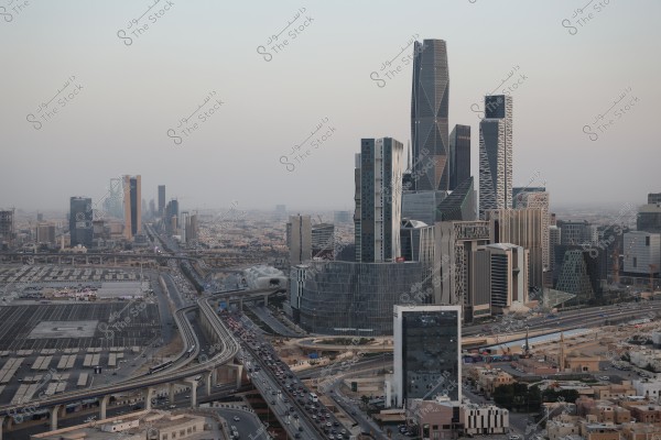 A view of Riyadh city in Saudi Arabia showcasing a collection of modern skyscrapers and notable architectural designs. The Kingdom Tower, a prominent landmark, is visible in the middle of the image. The streets are crowded with cars under a cloudy sky.