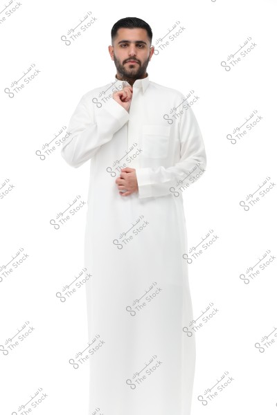 An image of a man standing wearing a traditional white garment known as a thobe. The man is set against a white background and has a calm expression on his face. The thobe is indicative of traditional attire commonly found in Gulf countries like Saudi Arabia.