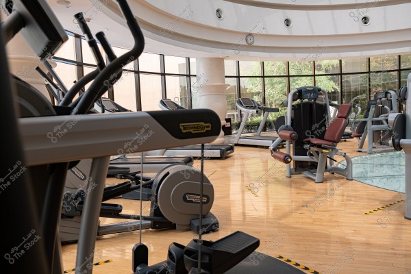 An image of a modern gym with various fitness equipment such as treadmills and other exercise machines. The floor is wooden, and the gym walls have large glass windows overlooking an area with green trees. The ceiling is circular, and there is a wall clock in the center.