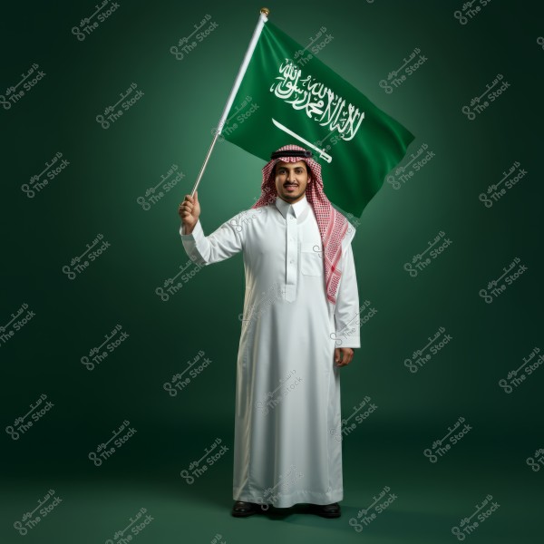 A photo of a man standing against a light green background, wearing a white thobe and a red and white shemagh. He is holding the Saudi Arabian flag in one hand. The image represents national pride and Saudi traditions.