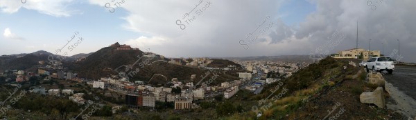 Panoramic view of a city in a mountainous area with multiple buildings spread across the hills, with a cloudy sky in the background. On the sloping road on the right of the image, two parked vehicles are visible.