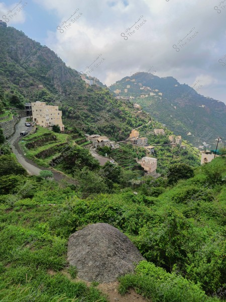 The image depicts a scenic view of a green mountainous village, with houses nestled among tall mountains covered with dense vegetation. A winding road passes through the village, with some buildings scattered along the mountainsides. The sky is overcast, suggesting a humid and rainy atmosphere.