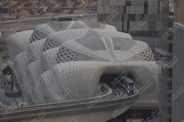 The image shows a modern train station with a contemporary and intricate architectural design in Saudi Arabia, featuring a high-speed train entering or leaving the station. The building has a wavy structure covered with geometric patterns, surrounded by towers with glass windows. In the background, a road is visible with cars moving around it.