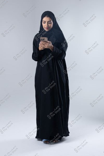 صورة لامرأة ترتدي عباءة سوداء وحجابًا, تقف وتنظر إلى هاتف محمول في يدها. تبدو في بيئة استوديو ذات خلفية رمادية محايدة. الأحذية ظاهرة قليلاً تحت العباءة.
