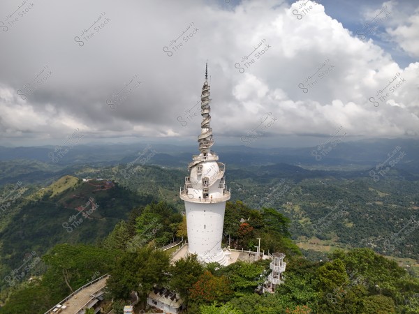 Image of a tall white communication tower located on the top of a mountain covered with green trees. The tower features multiple tiers and a circular base with a spiral mast at the top. The background shows dense clouds and an overcast sky, with a panoramic view of hills and a sprawling valley.