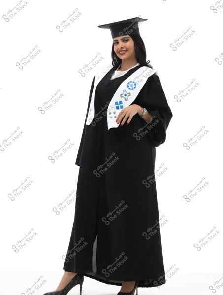 Image of a woman wearing a black graduation gown and a mortarboard. She has a white stole decorated with blue and grey patterns. The woman is standing with a smile on her face and wearing high-heeled shoes. The photo appears to be taken in a graduation celebration context.