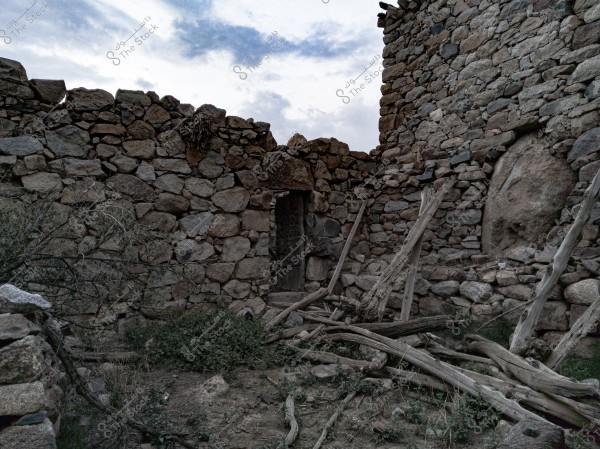 An image depicting the remains of an old stone structure in a desert area under a cloudy sky. The walls are made of irregular stones with a small doorway. There are worn wooden planks and dry branches scattered on the dirt ground, with some dry plants growing between the stones.