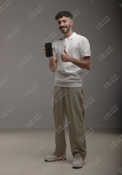 A man stands in a studio wearing a white polo shirt and beige trousers. He is holding a mobile phone in one hand and showing a thumbs-up gesture with the other. The background is plain gray.