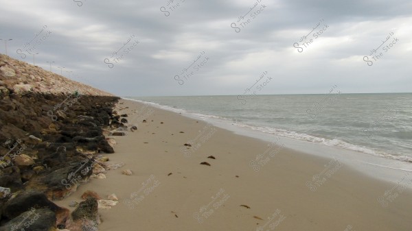 An image of an empty beach stretching to the horizon under a cloudy sky. The left side of the image features a collection of large rocks piled alongside the beach, while gentle waves touch the sand on the right side.