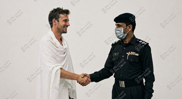 The image shows two men shaking hands. The man on the left is wearing a white ihram, indicating he is on pilgrimage, and is smiling. The man on the right is wearing a black military uniform with a cap and a face mask, suggesting he is from Saudi Arabia. The background is completely white.