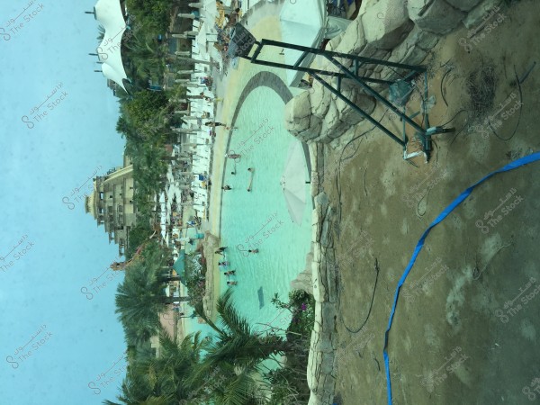 An image of a large outdoor water facility featuring a wide circular pool surrounded by white lounge chairs and umbrellas. Several people are visible in and around the pool. The pool area is bordered by palm trees, and a large building is in the background, suggesting a tropical or resort atmosphere.