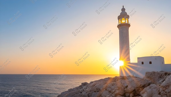 The image shows a sunset behind a white brick lighthouse, with the sun\'s rays shining vividly through the lighthouse arches. The calm blue sea is in the background, while the sky displays a gradient of warm colors between orange and blue. White rocks are piled in the foreground.