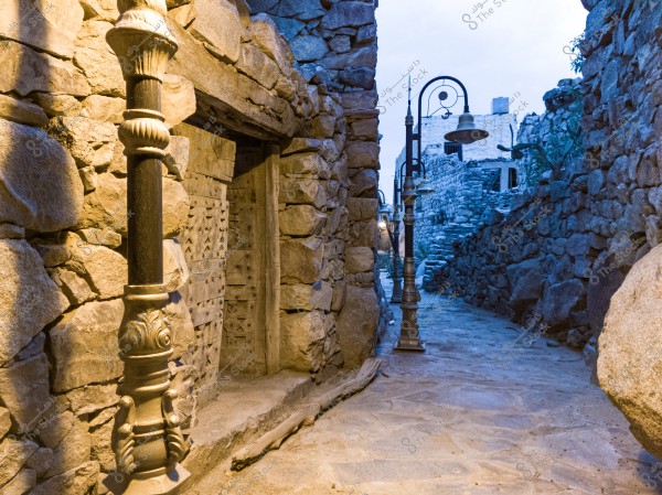 An image of a narrow alleyway between stone walls in an old village, featuring traditional lamps hanging from metal posts along the path. Intricately carved wooden doors are visible on the left side, and the atmosphere appears cold and slightly dim with a cloudy sky.