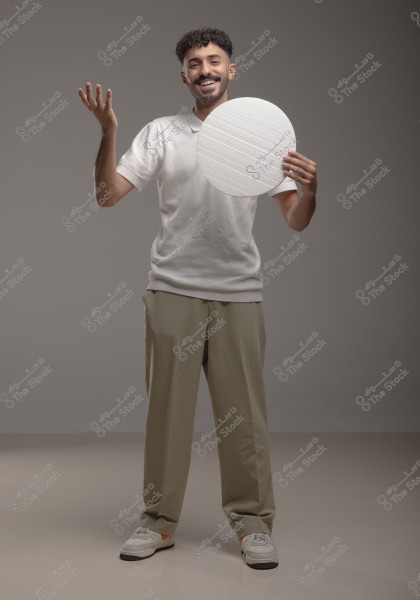 A smiling man with short curly hair wearing a white shirt and beige trousers stands against a gray background, raising his left hand in the air while holding a white circular object.