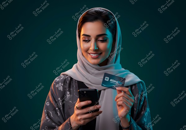 A portrait of a woman wearing a light gray hijab and a black abaya with silver patterns, holding a payment card labeled \"AlimaPay\" along with her mobile phone. The background is dark, and the woman is smiling.