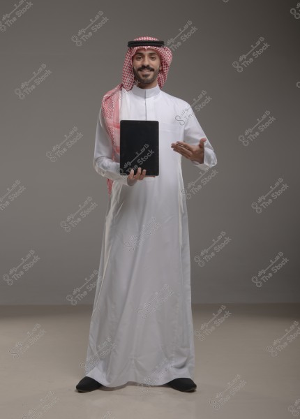 A portrait of a man standing in a studio against a grey background, wearing a white thobe, a red and white checkered ghutra, and a black agal. The man is holding a tablet in one hand and gesturing with the other. The outfit suggests traditional Gulf attire, possibly from Saudi Arabia.