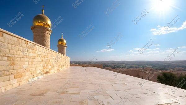 The image shows a beautiful architectural structure featuring minarets with golden domes. In front of it, there is a large expanse of light-colored stone flooring. The sky is clear and blue, with the sun in the upper right corner, creating a striking contrast with the stone architecture. In the background, a city stretches out to the horizon, surrounded by green natural landscapes.
