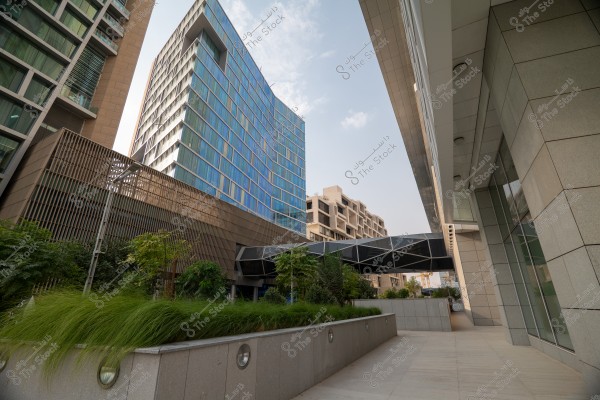 An image of a modern architectural facade featuring tall buildings with glass and metallic exteriors. Centrally positioned is a blue glass building with intricate geometric designs, surrounded by green spaces and trees, with a covered walkway connecting to another building on the right. The sky is clear with a few scattered white clouds.