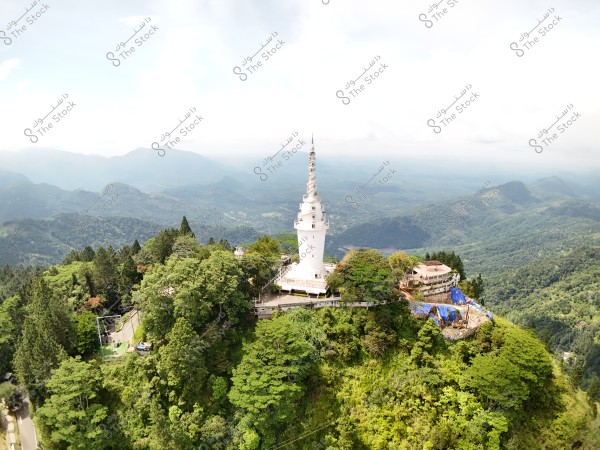 Aerial view of a tall white tower situated in a green mountainous area covered with dense trees. The tower is surrounded by several small buildings and is perched on top of a hill. Mountains and valleys are visible in the background under partially cloudy skies.